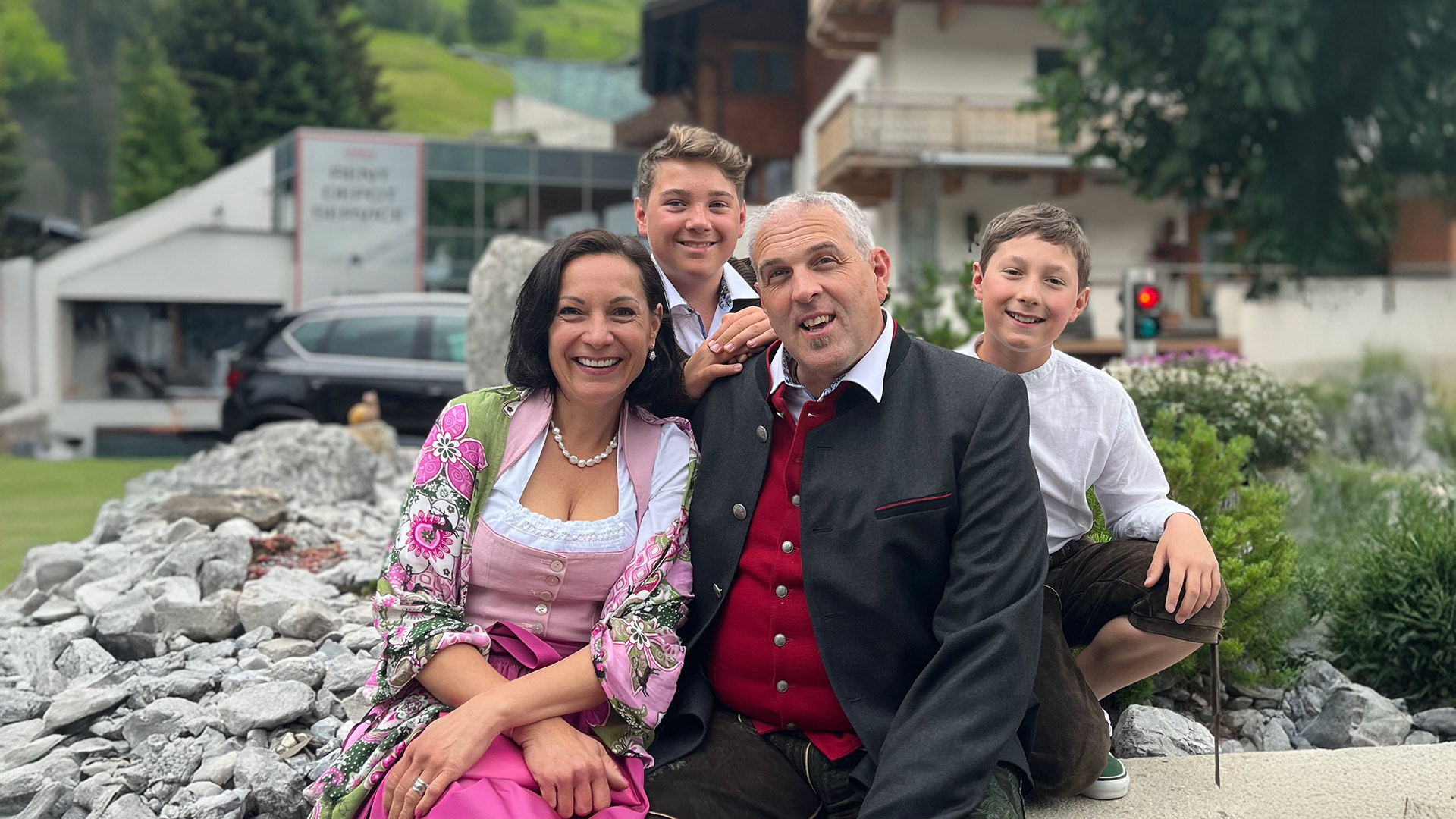 Familie Weiskopf in traditioneller Kleidung sitzt draußen vor einem Steingarten, mit Alpenhäusern im Hintergrund.
