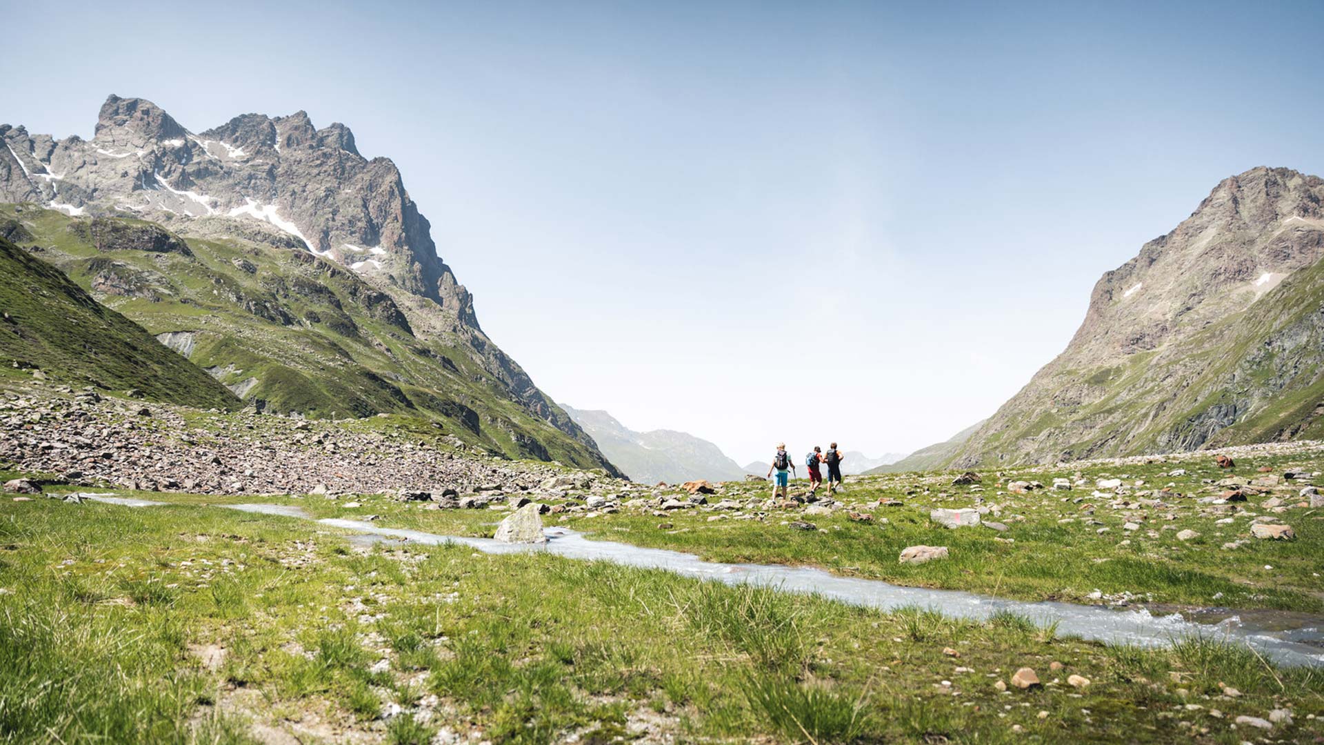 Wanderer in weitem Tal mit Bachlauf, umgeben von grünen Hängen und steilen Felsgipfeln bei klarem Sommerwetter.