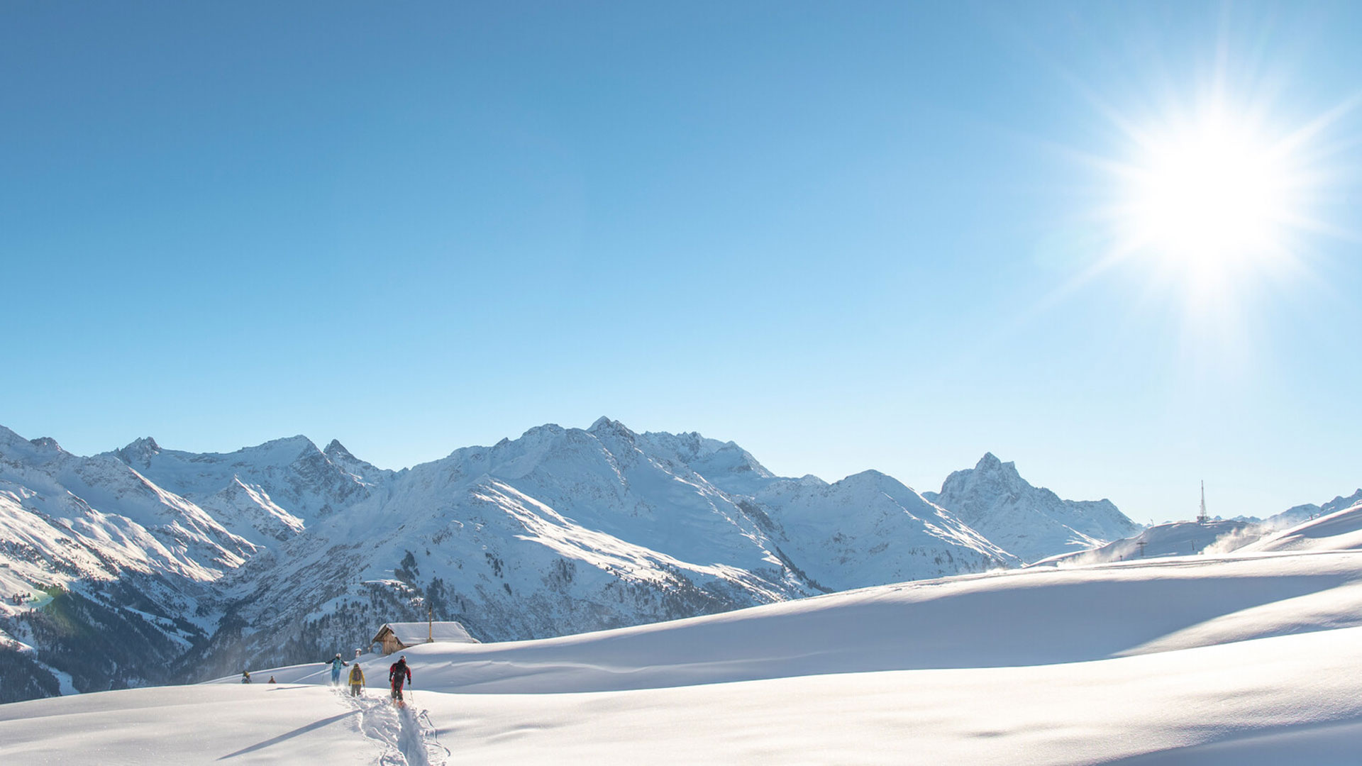 Schneeschuhwanderer in unberührter Winterlandschaft unter strahlender Sonne mit Panorama auf verschneite Alpengipfel.
